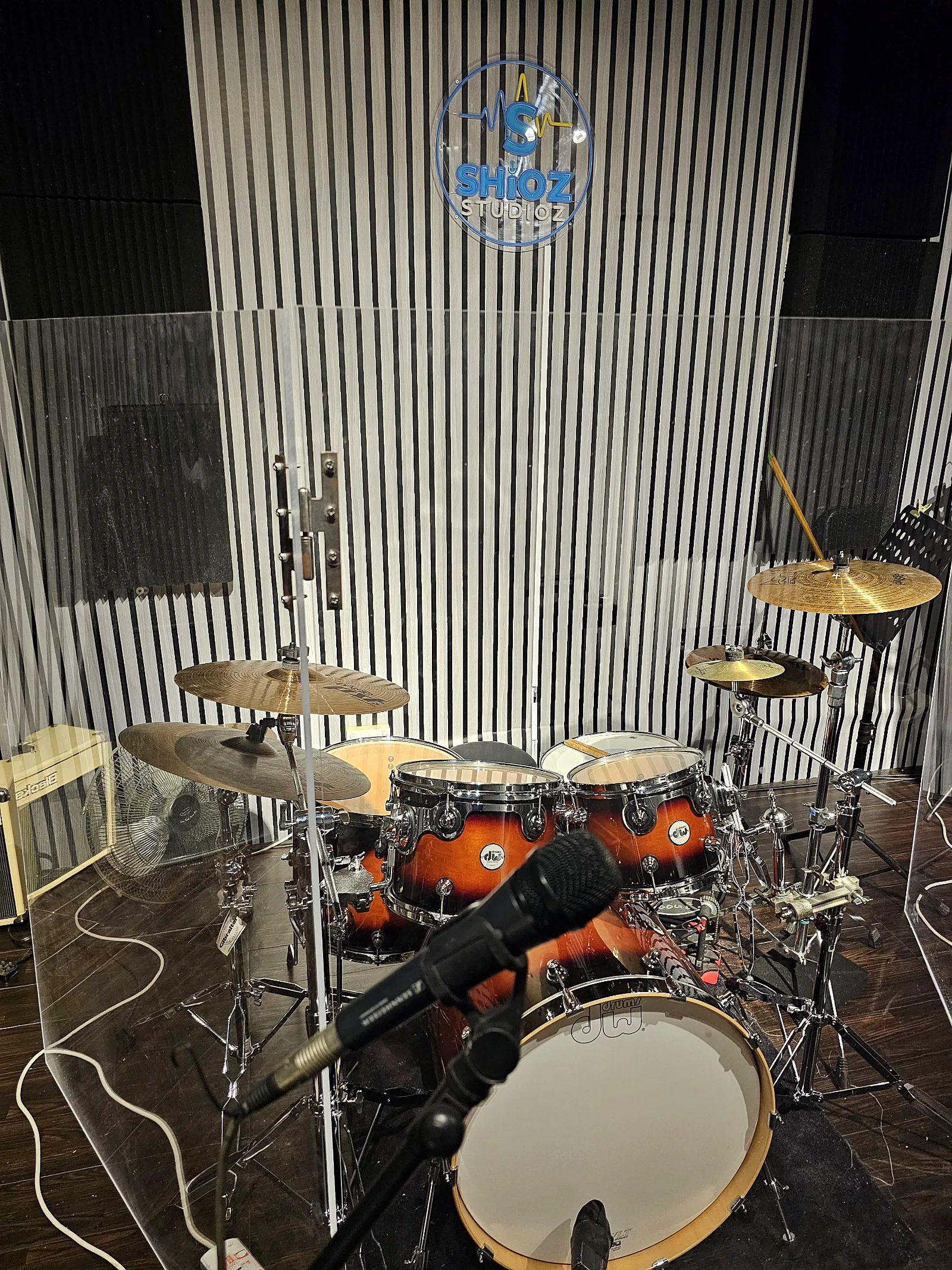 DW drum kit inside the clear drum shield under the neon Shiokz sign
